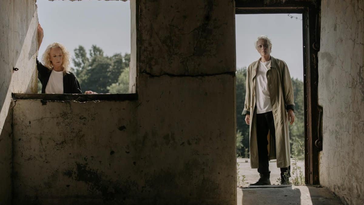 An elderly couple stands in an old building, surrounded by light and shadow, evoking themes of aging and nostalgia.