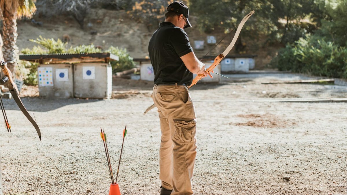 An adult male practicing archery outdoors with a recurve bow, focused on target practice.