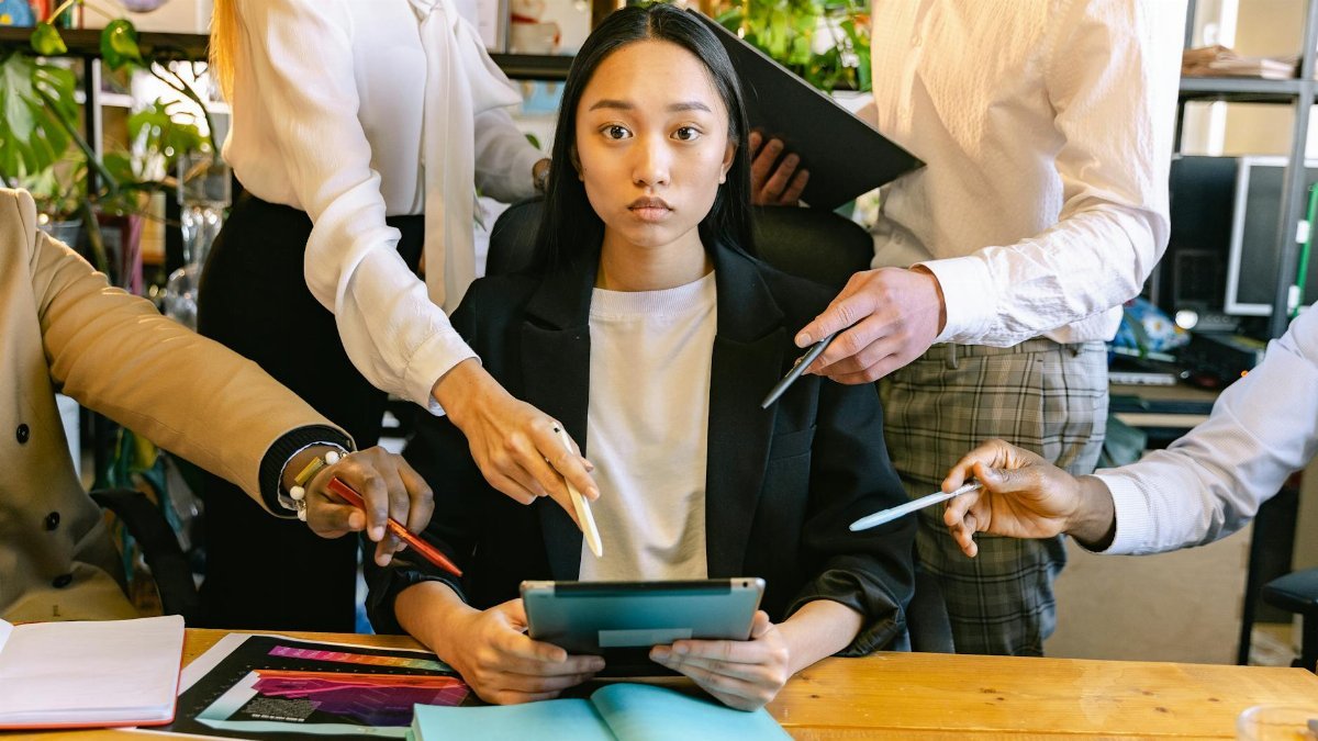 An overwhelmed office worker surrounded by colleagues requesting her attention during a busy workday.