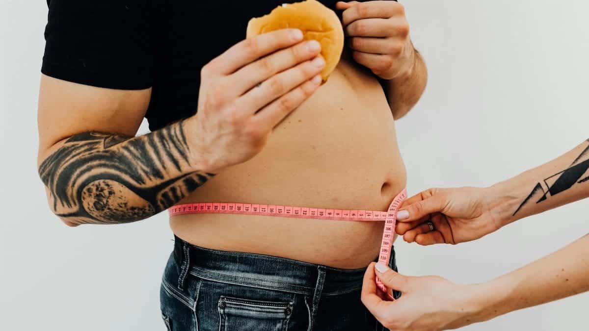 A man with tattoos holding a burger as another person measures his waistline with a tape measure.