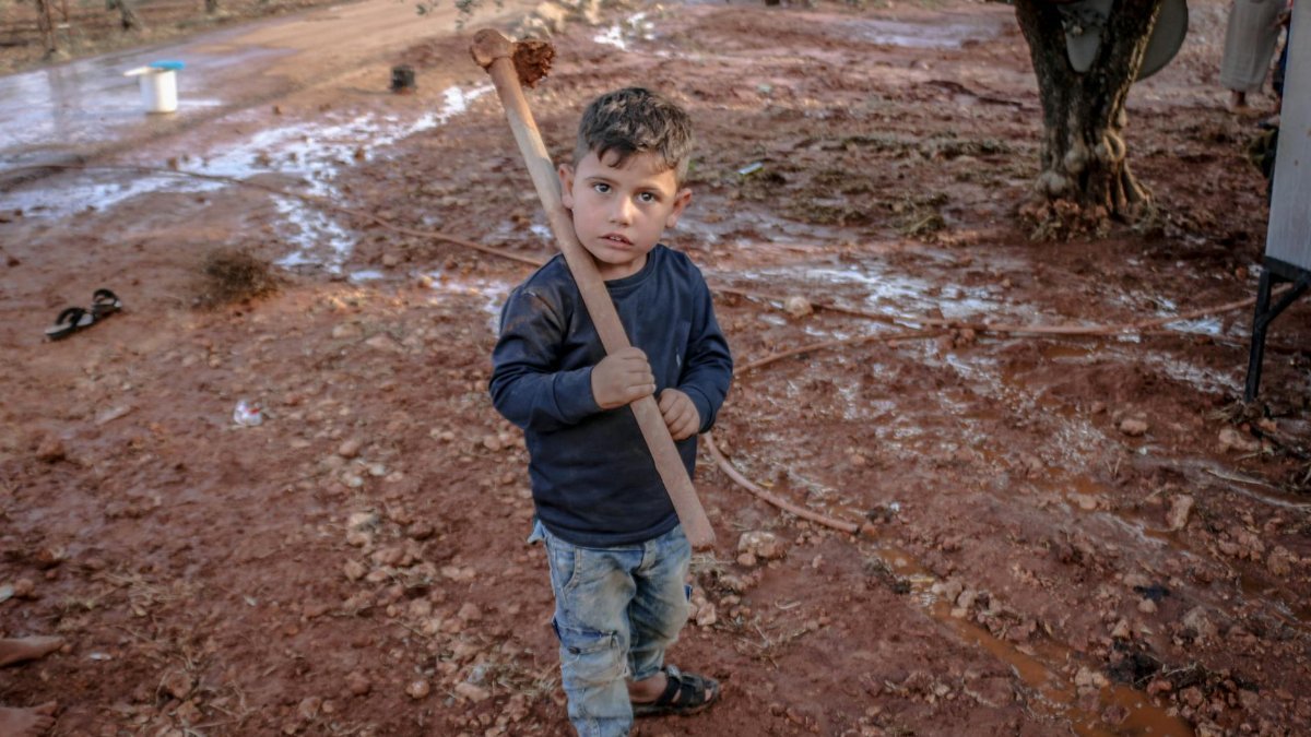 A young boy holding a stick stands on muddy ground in rural Idlib, Syria.
