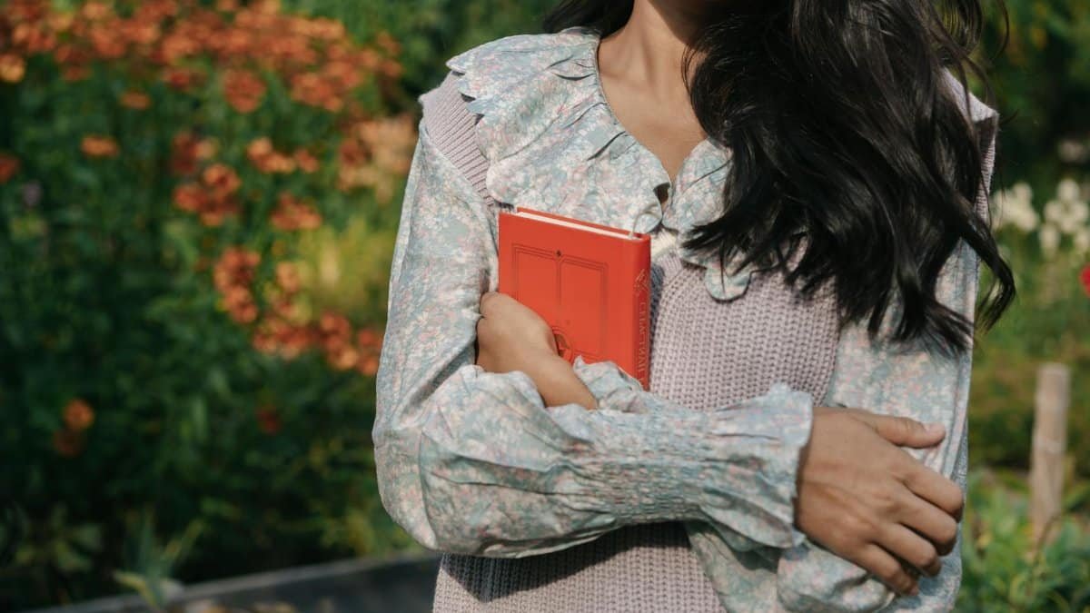 A woman stands in a garden, arms crossed holding a red book, wearing a floral top with long sleeves.
