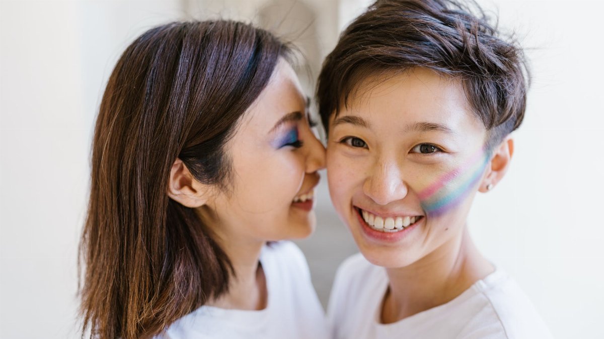 A joyful portrait of two women with rainbow face paint, embracing and smiling indoors.