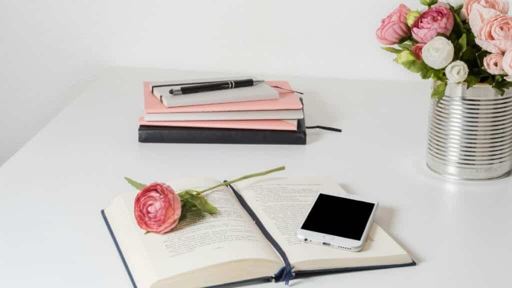 A stylish home office setup with open book, smartphone, and flowers on a white desk.