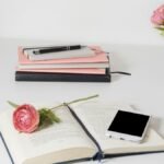A stylish home office setup with open book, smartphone, and flowers on a white desk.