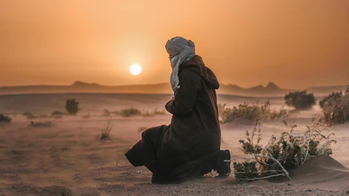 A man kneels in the desert at sunset, wearing traditional clothing and a turban.