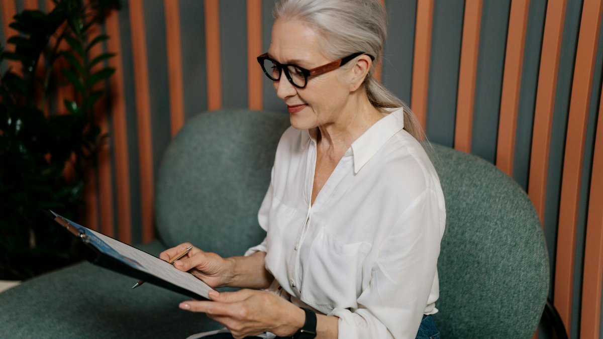 Elderly woman in glasses reviewing documents on clipboard, sitting indoors with a focused expression.