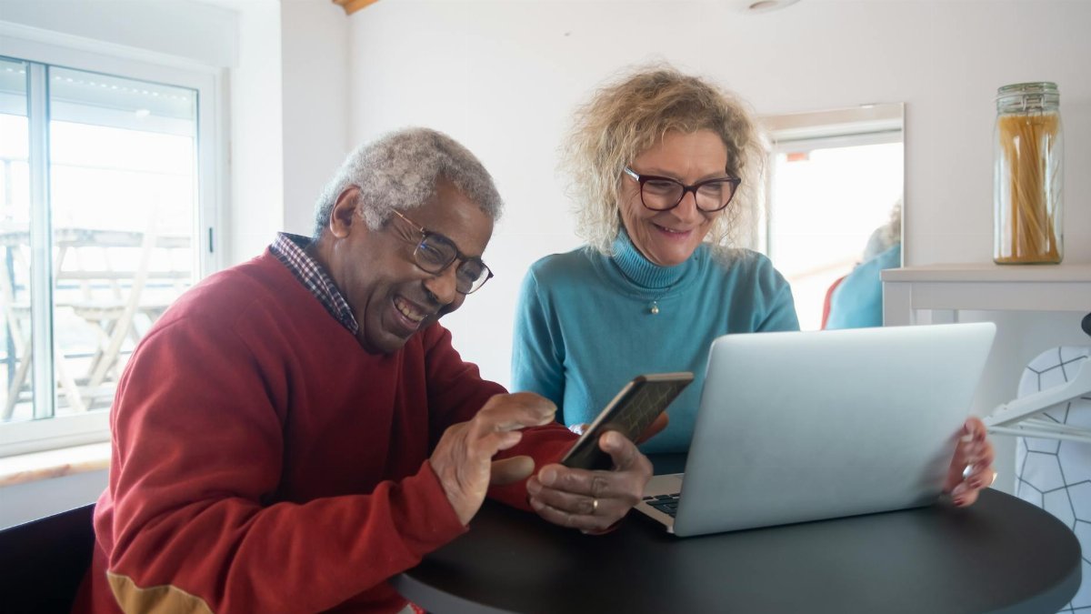 Smiling elderly couple using a smartphone and laptop indoors, enjoying online activities.