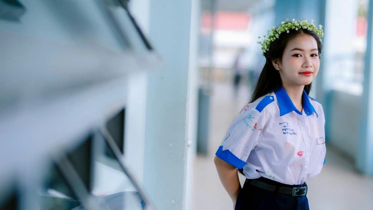 A young woman in a uniform with a floral crown stands indoors, smiling softly.