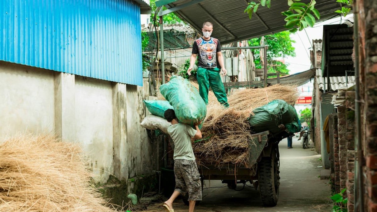 Two men load straw bundles onto a cart in a rural alley, working hard on a bright day.