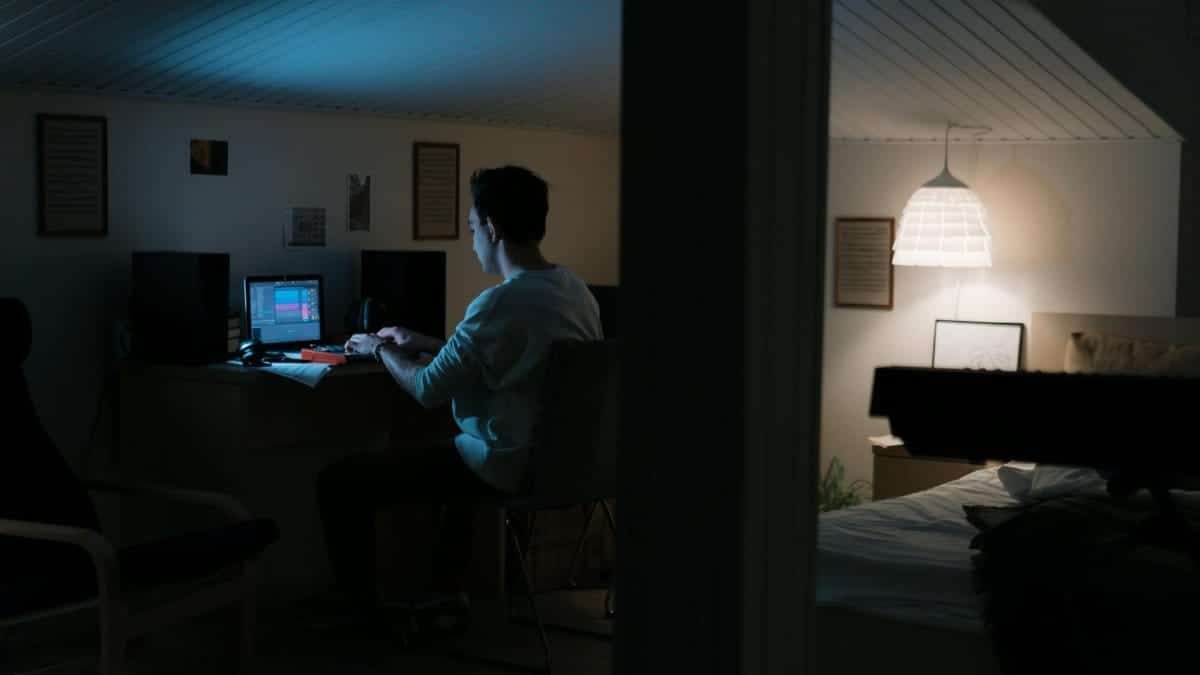 A man working at his desk in a dimly lit bedroom, using a computer with digital audio software.