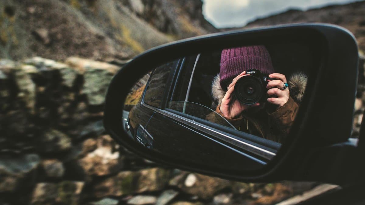 Reflective photography of a person with camera in car side mirror outdoors.