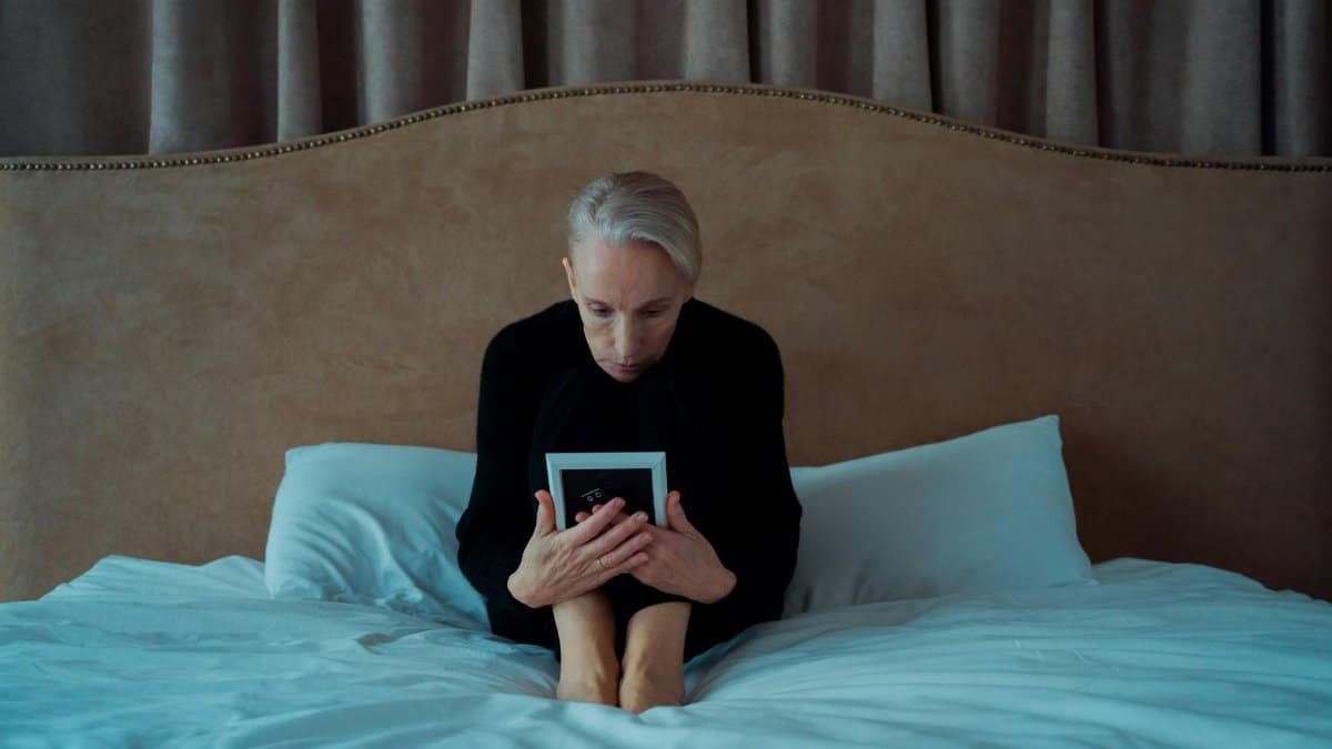 An older woman sits on a bed holding a photo, capturing a moment of reflection.