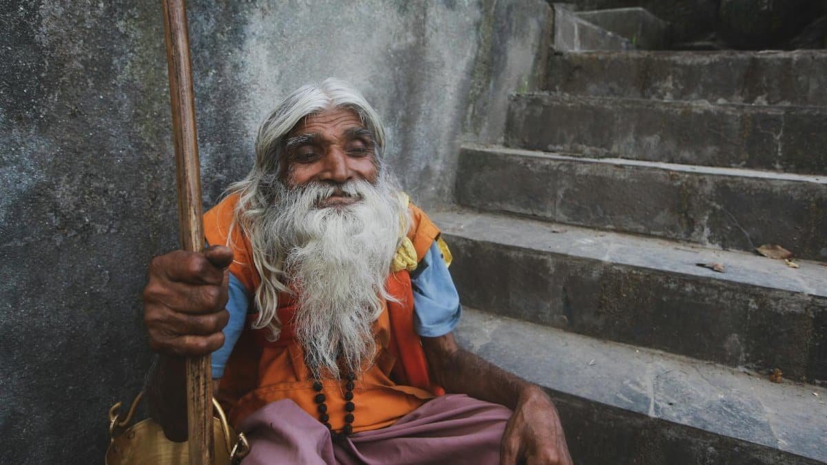 Portrait of an elderly man with long white beard and colorful attire, sitting on stone stairs.