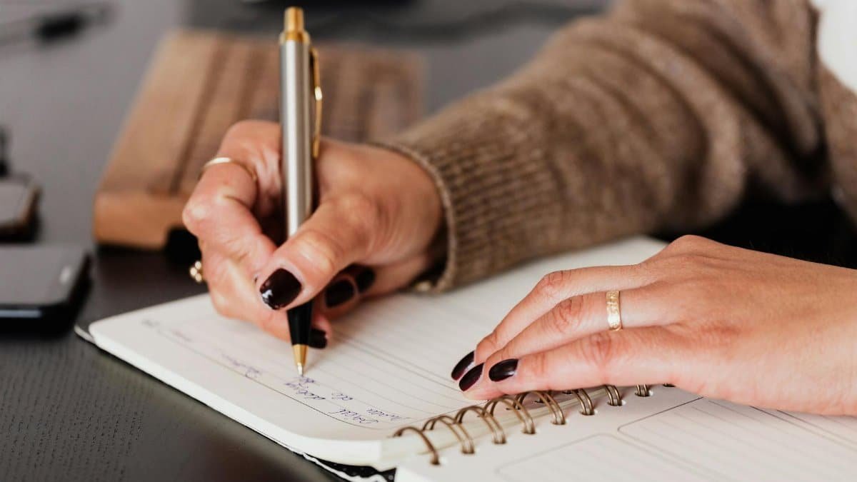 Crop unrecognizable female with stylish manicure sitting at black desk with keyboard and smartphone and taking notes with silver pen in notepad