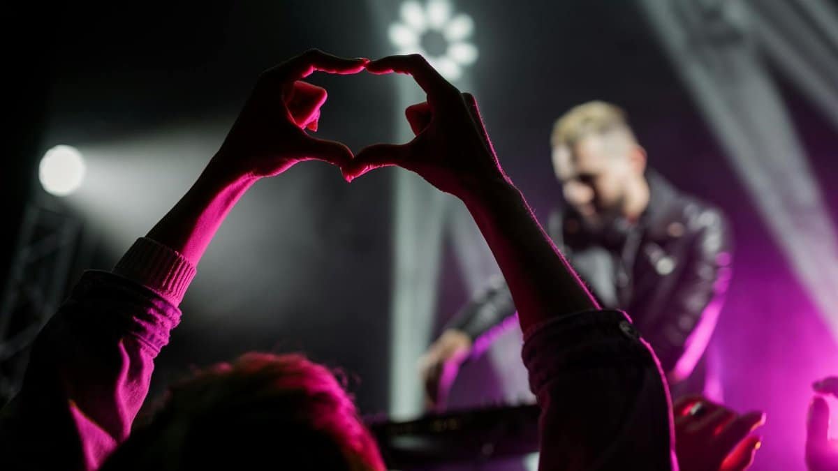 A lively concert scene featuring a heart-shaped hand gesture against a vivid purple backdrop.