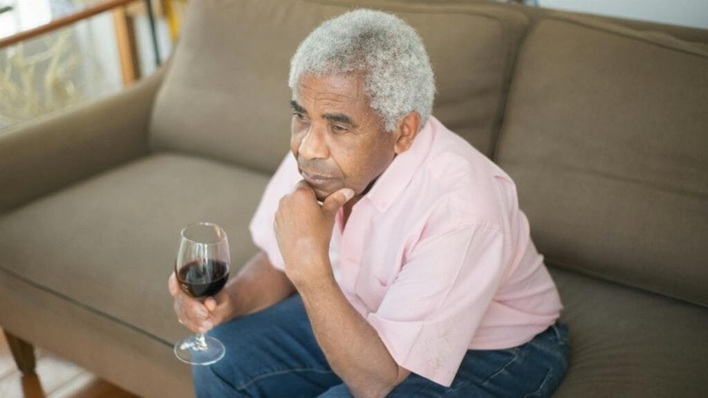 Contemplative elderly man sitting on a sofa holding a glass of wine indoors.