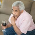 Contemplative elderly man sitting on a sofa holding a glass of wine indoors.