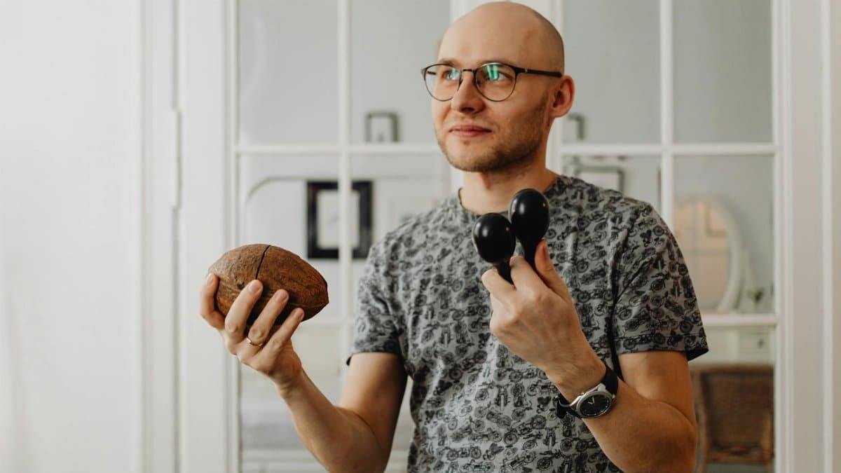 Bald man with eyeglasses holding maracas and coconut shell indoors.