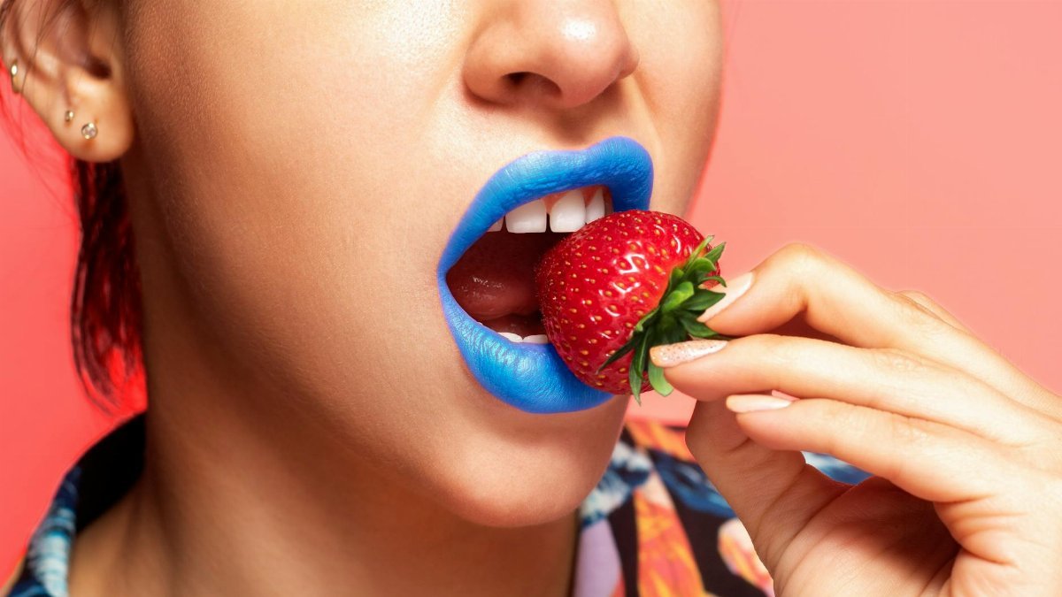 Close-up portrait of a woman with blue lipstick holding a fresh strawberry.