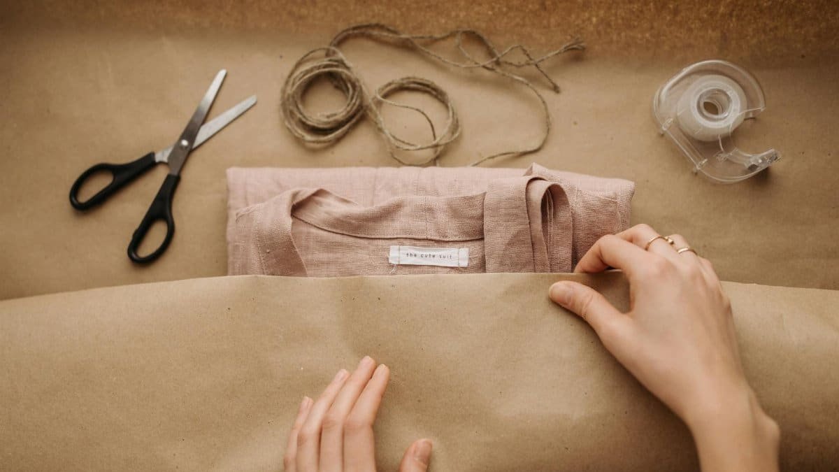 Close-up of hands wrapping a linen shirt with twine, scissors, and tape on brown paper.