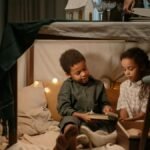 Two children enjoy reading together inside a cozy fort under a table with festive decorations.