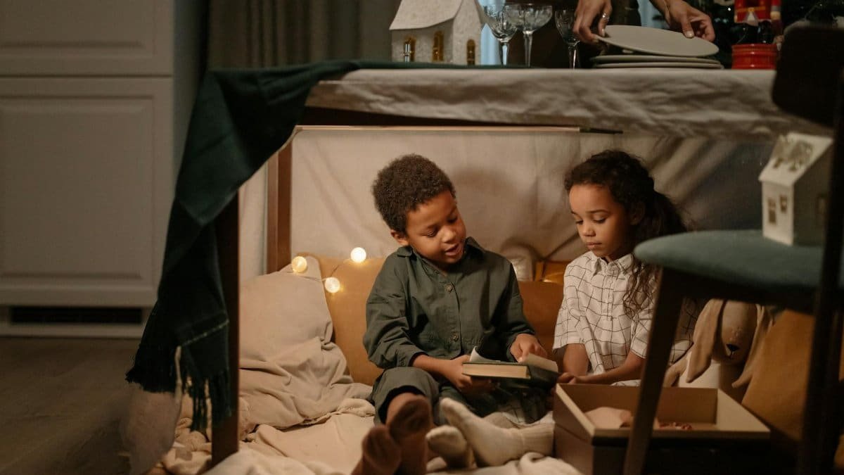 Two children enjoy reading together inside a cozy fort under a table with festive decorations.