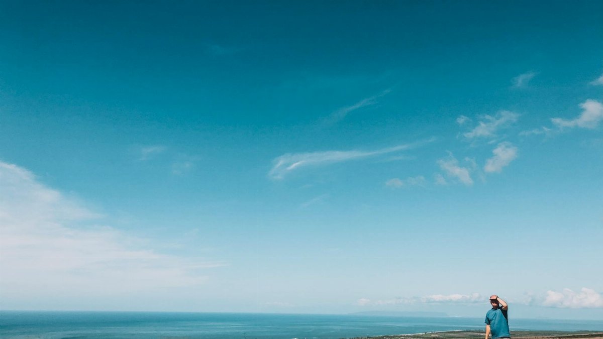 A man standing in a field overlooking a vast ocean and clear blue sky, enjoying the summer atmosphere.