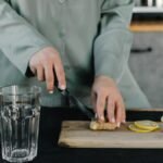 Hands cutting ginger next to lemons, a glass, and water bottle on a wooden board indoors.