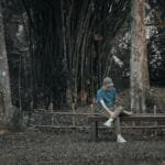 A young man sits on a bench in a peaceful forest setting in Thailand, engrossed in his phone.