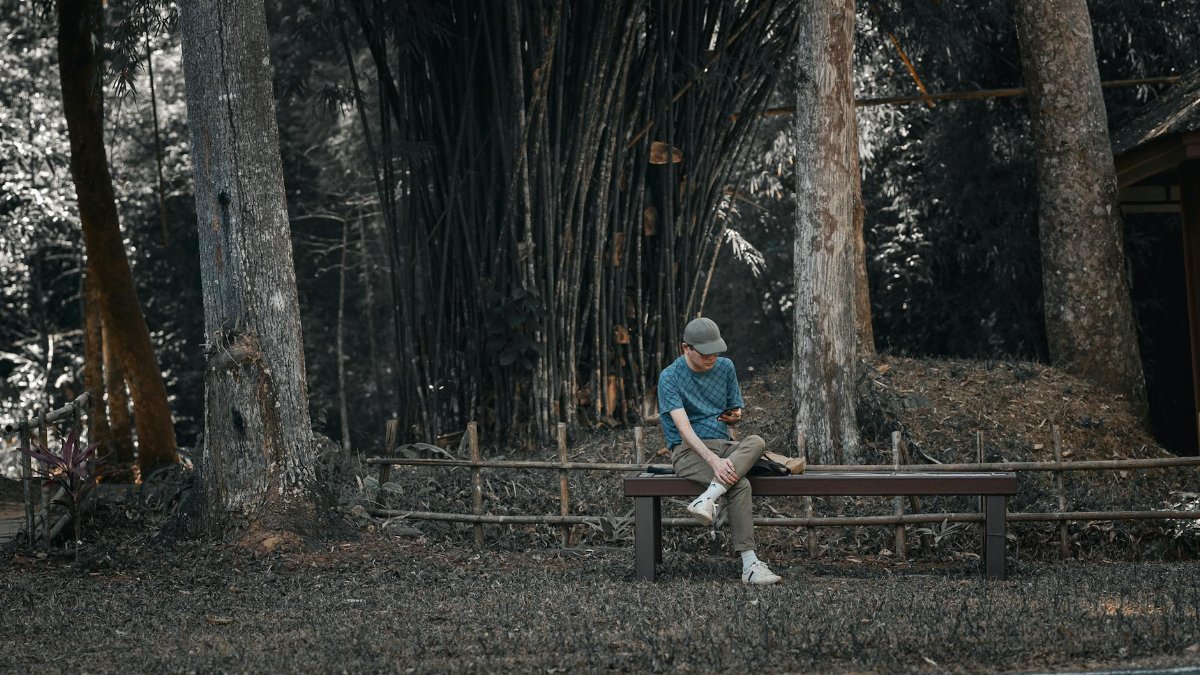 A young man sits on a bench in a peaceful forest setting in Thailand, engrossed in his phone.