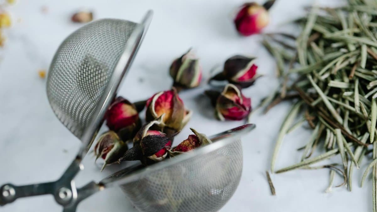 Close-up of rosebuds and herbs in a stainless steel tea strainer on a white surface.