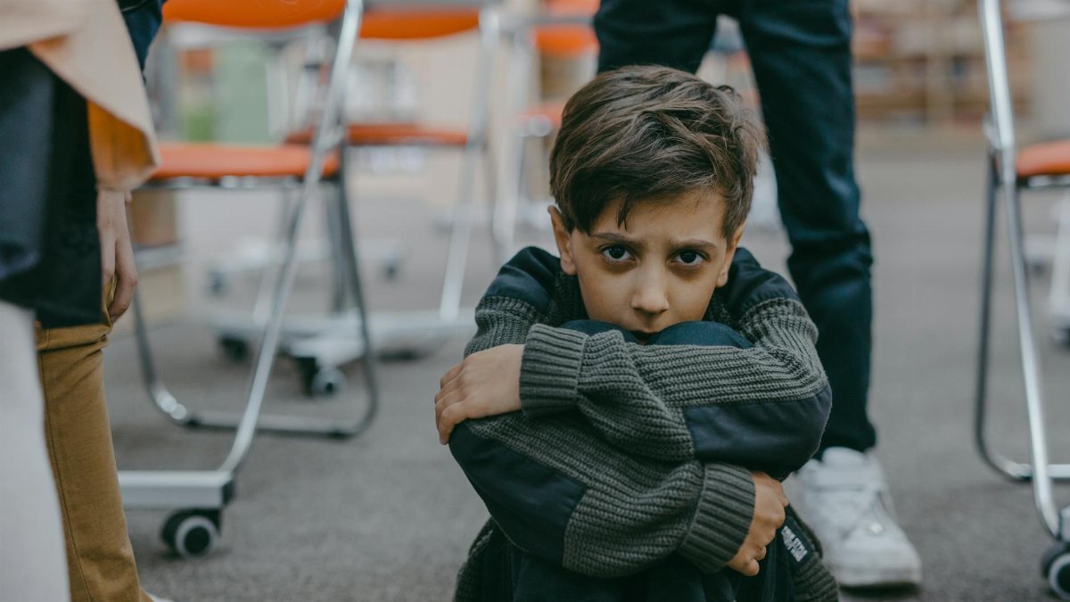 A young boy sits on the floor of a classroom, expressing fear and vulnerability.