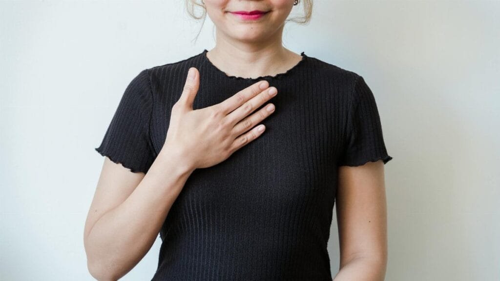 Asian woman in a black shirt expressing gratitude with a hand gesture against a neutral background.
