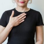 Asian woman in a black shirt expressing gratitude with a hand gesture against a neutral background.