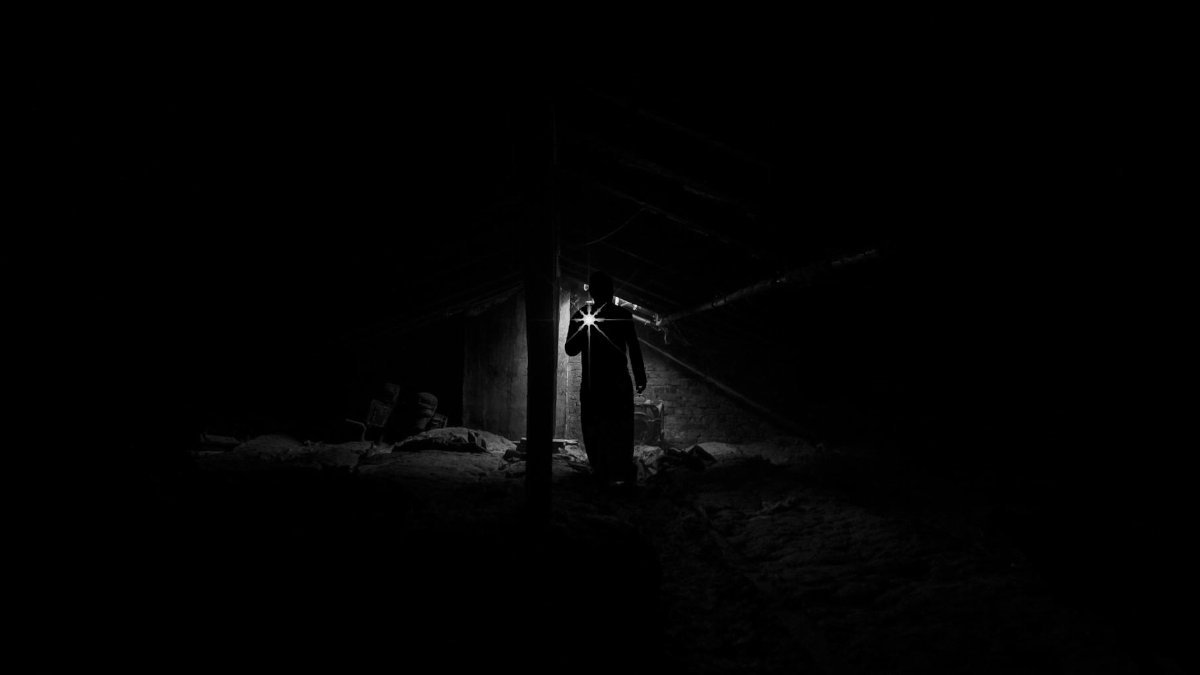 A shadowy figure explores a dimly lit, eerie attic with a flashlight.
