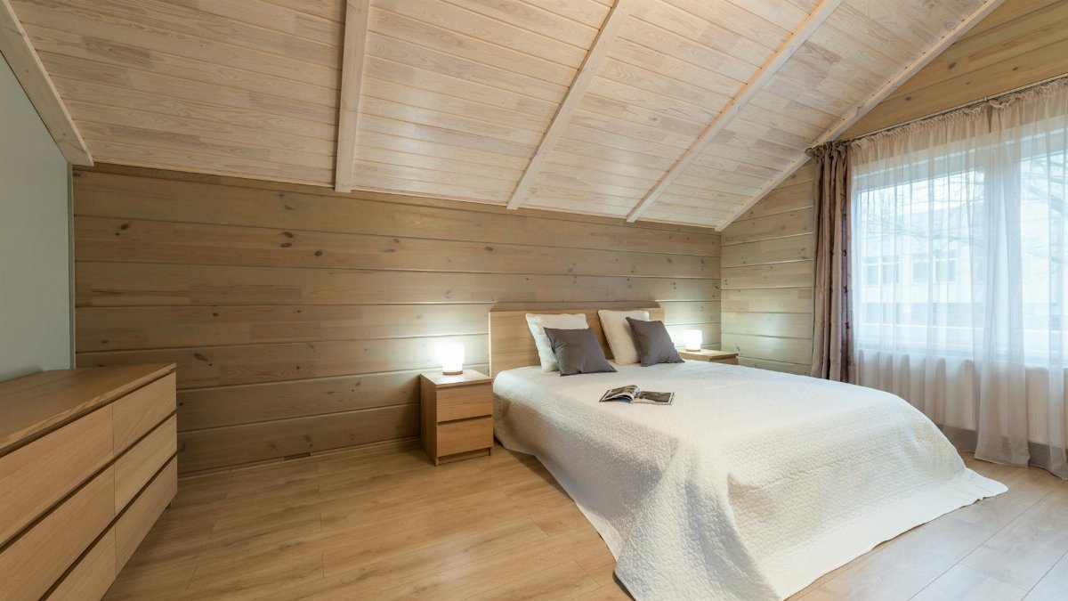 Comfortable bed with pillows and opened magazine with luminous lamps on nightstands on wooden mansard of cottage