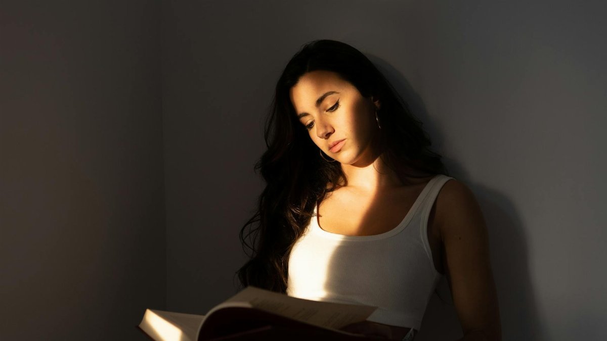 Young woman in white top reading a book, highlighting the interplay of light and shadow indoors.