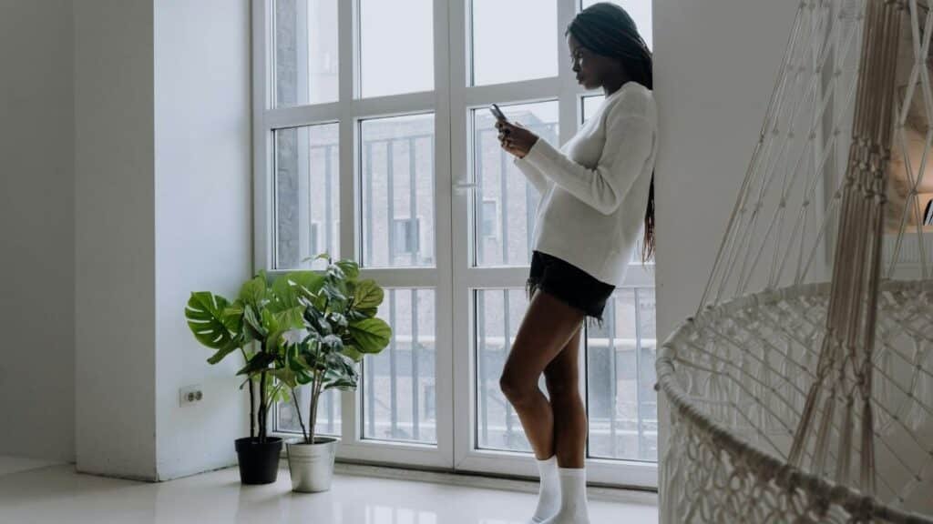 A young woman with braids uses her mobile phone in a minimalist room with plants and a hammock chair.