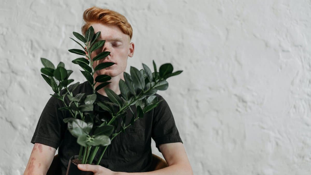 Red-haired young man in black t-shirt holding a plant indoors with eyes closed against white background.