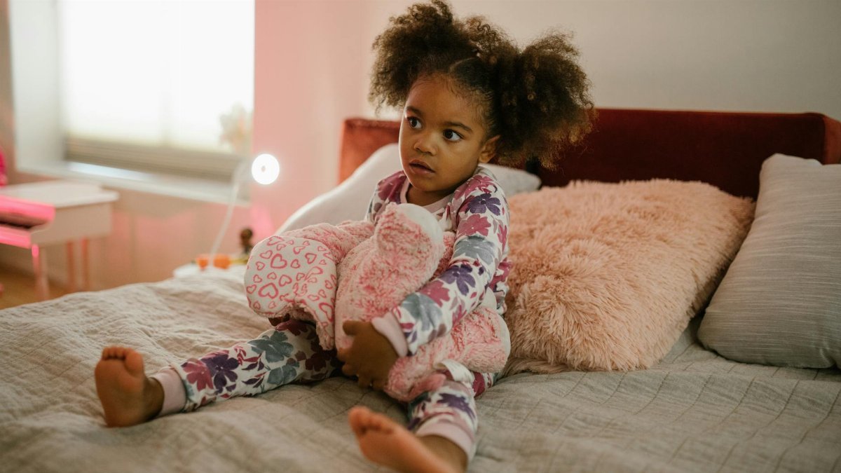 A young girl in pajamas sits on her bed holding a stuffed animal in a cozy bedroom.