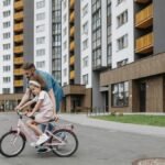 Father guiding daughter on a bike ride outside modern apartments. Bonding and teaching moment.