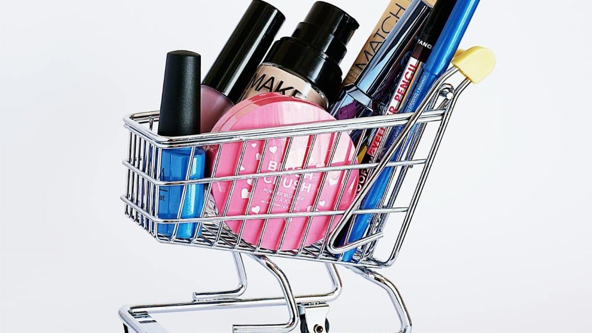 Miniature shopping cart filled with a variety of cosmetics on a white background.