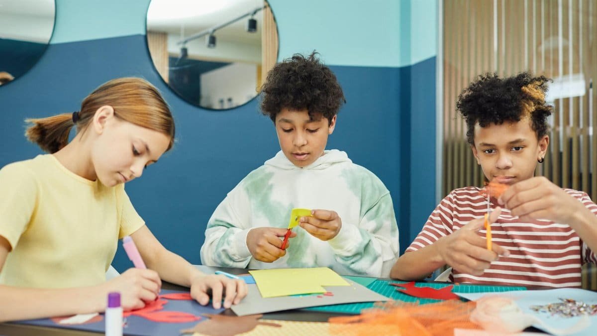 Group of concentrated multiracial children making artworks with colorful paper and scissors while sitting at table in light room