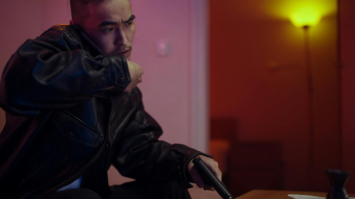Man in a black leather jacket holding a gun while making a phone call indoors.