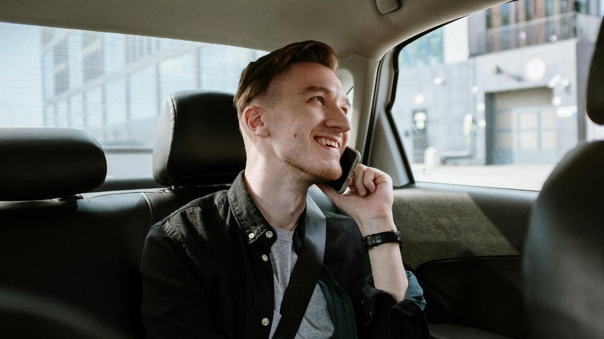Smiling young man enjoying a phone call while seated in the back seat of a car during a daytime ride.
