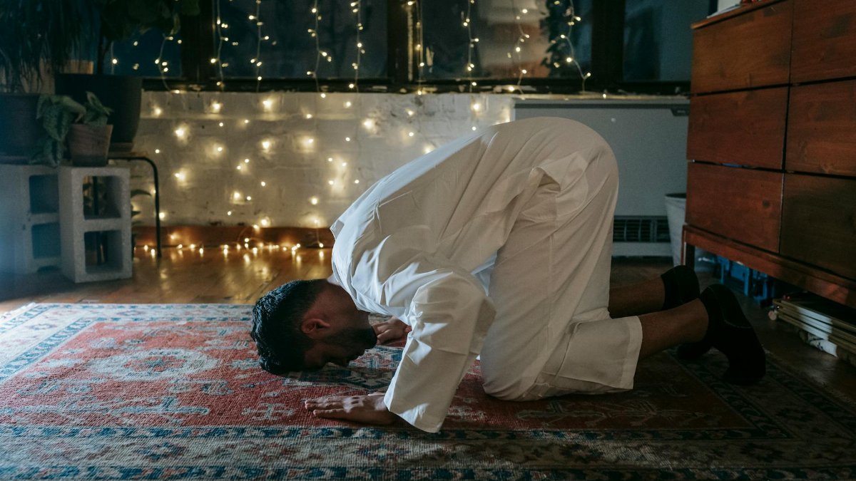 A Muslim man in traditional attire praying on a rug indoors with ambient lighting.