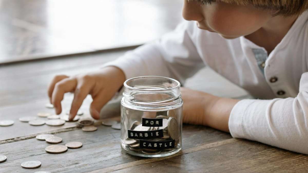 A young child collects coins in a jar labeled 'For Barbie Castle', symbolizing saving and dreams.