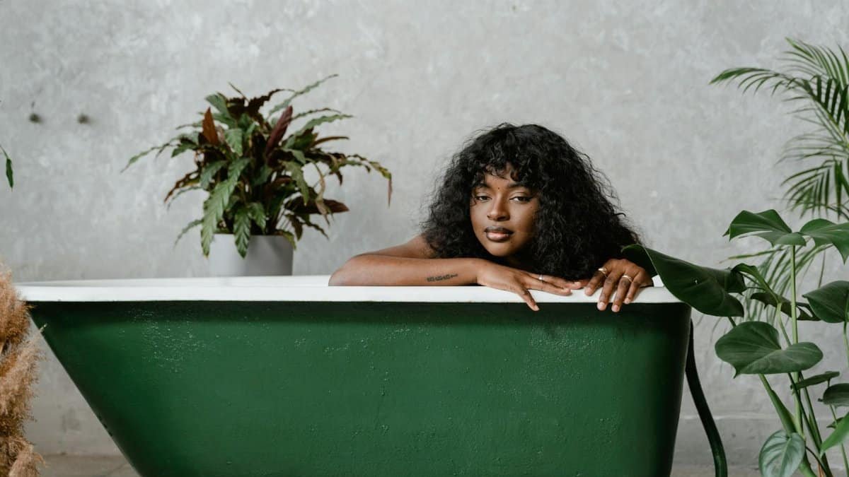 A woman with curly hair relaxes in a green bathtub, surrounded by potted plants against a concrete wall.