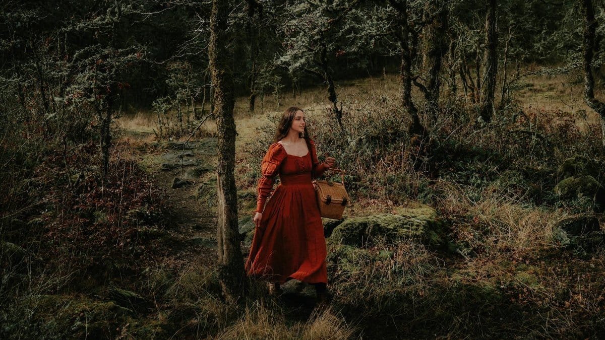 A woman in a red gown carries a wicker basket while strolling through a lush forest in Portland.